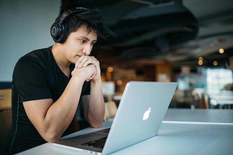 A young man sitting at a desk, looking at a laptop and listening to audio through headphones.