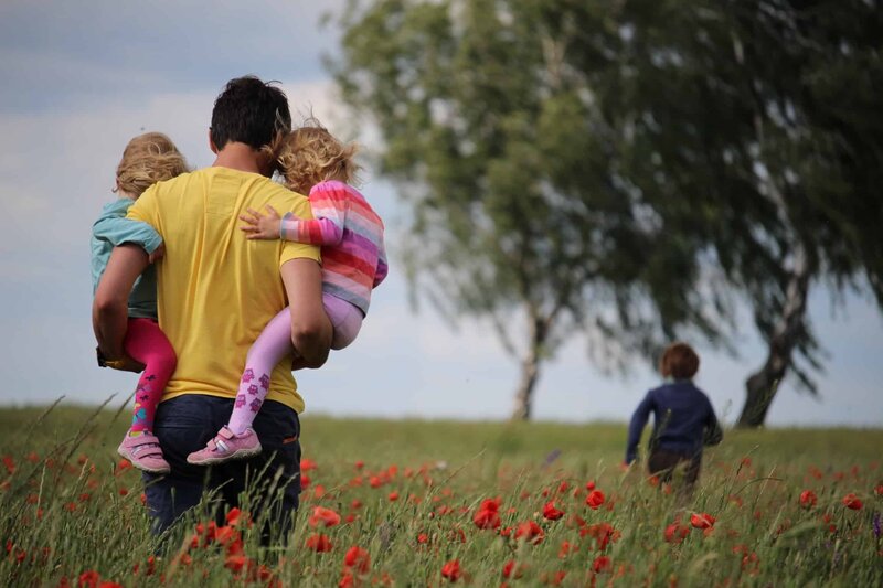 A photo of a man carrying 2 small children, one on each hip. He is walking through a field of poppies towards 2 trees on a sunny Summers day. There is another figure nearer the trees. The photo is taken from the back.
