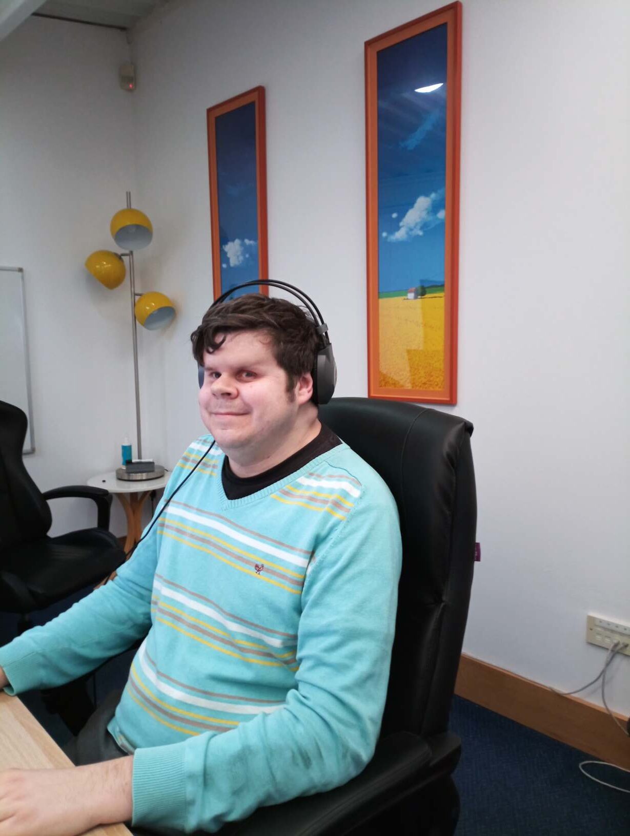 Dave sitting at his desk, smiling, in A2i's office. He is wearing a turquoise jumper and has headphones on
