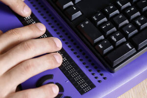 A refreshable Braille display next to a computer keyboard. The photos also shows part of someone's hands, who is using the refreshable Braille display.