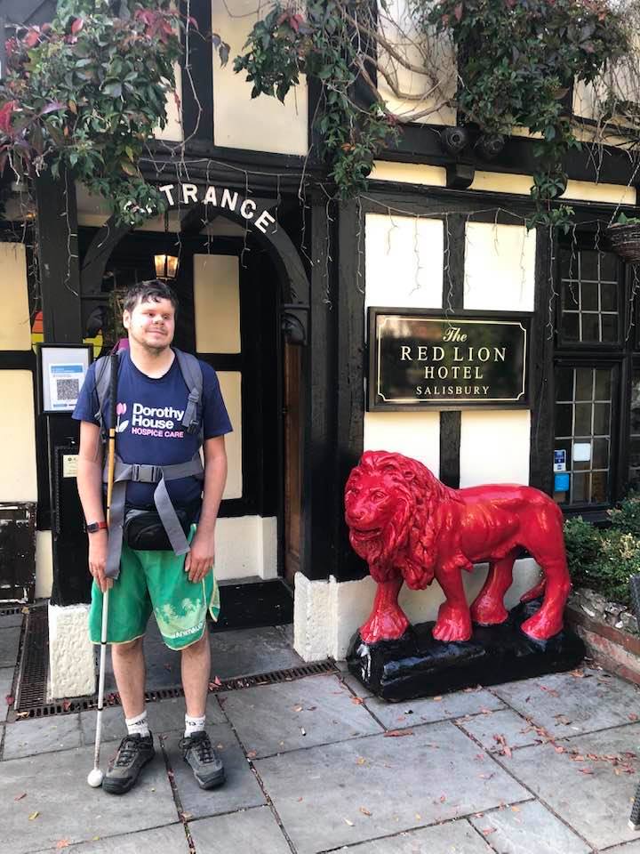 Dave standing outside the Red Lion pub in Salisbury, at the end of one of his charity walks. Dave is wearing shorts, a t-shirt and a big rucksack! The pub is 800 years old, with pretty hanging baskets and a statue of a red lion, next to Dave.