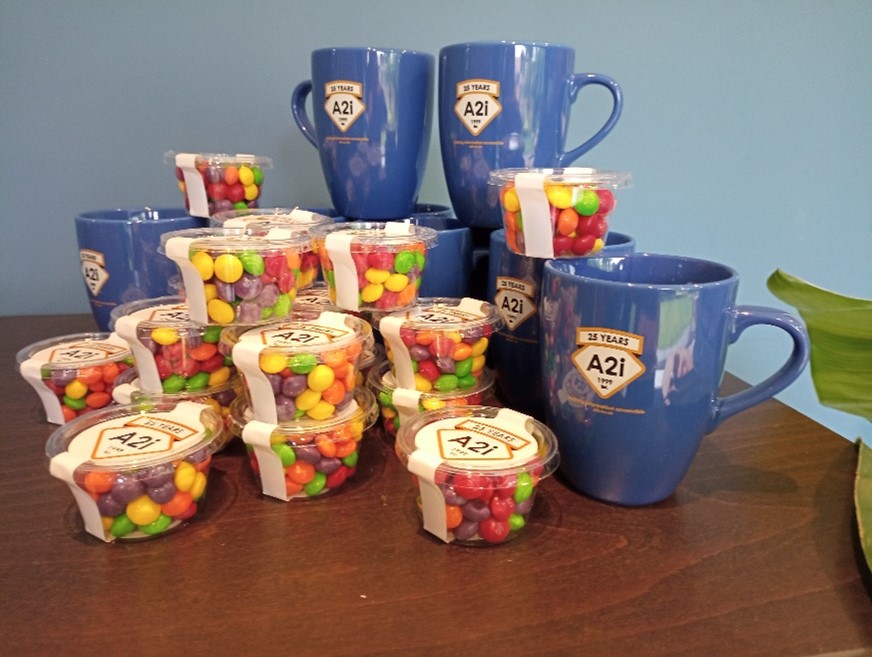 Blue mugs with A2i's logo, and mini pots of Skittles sweets, with packaging that includes A2i's logo.