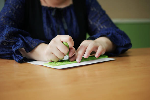 Close-up of blind woman using a slate and stylus to write Braille. The stylus is a wooden handle with a metal point sticking out that is used to press dots into the paper that is under the slate. The slate is green plastic and is used to guide the point of the stylus.