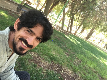 Rick, sitting in a park, smiling. Rick has black hair, a moustache and a small beard, and he's wearing a pale green jumper.