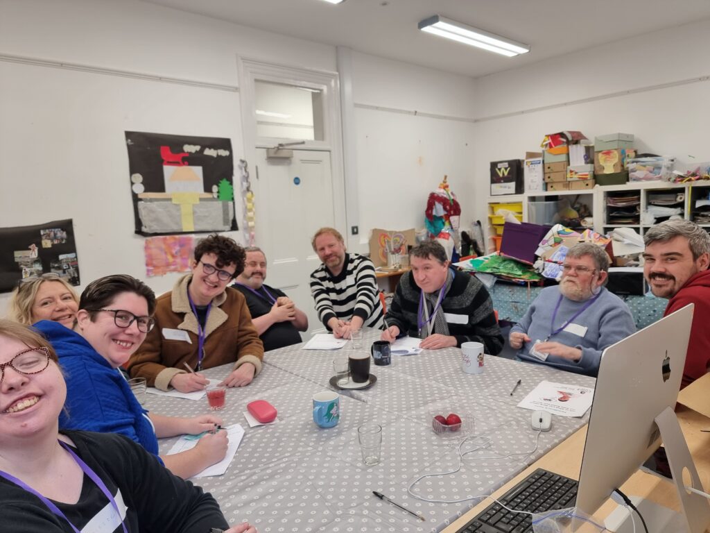 8 adults with a learning disability, and 1 A2i staff member, sitting around a table together and smiling for the camera.