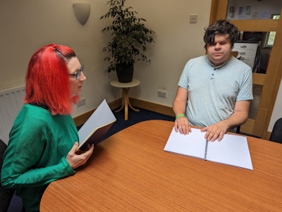 Susie (employer) and Dave (employee) sitting at a meeting table together. Susie is reading from a booklet, Dave if reading a Braille document.