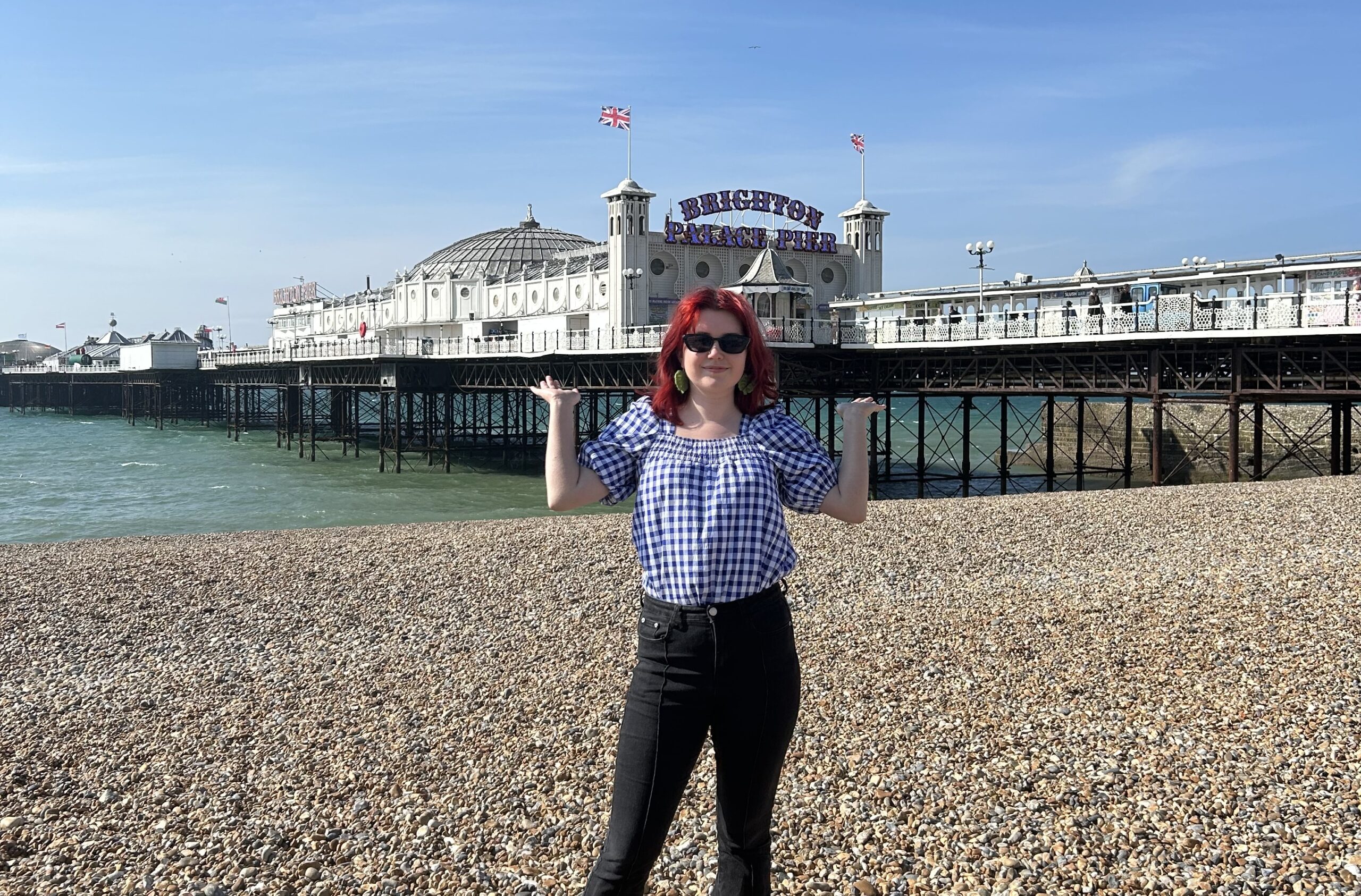 A photo of Elle on a beach on a sunny day, with a big pier in the background. Elle is wearing black jeans and a blue and white checked shirt, and sunglasses. She is smiling at the camera
