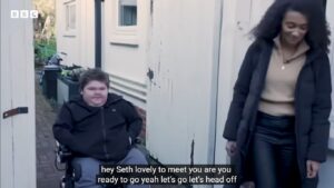 Screen shot of a BBC documentary showing the presenter with a disabled child in a wheel chair. The content of their conversation is shown as subtitles at the bottom of the screen 
