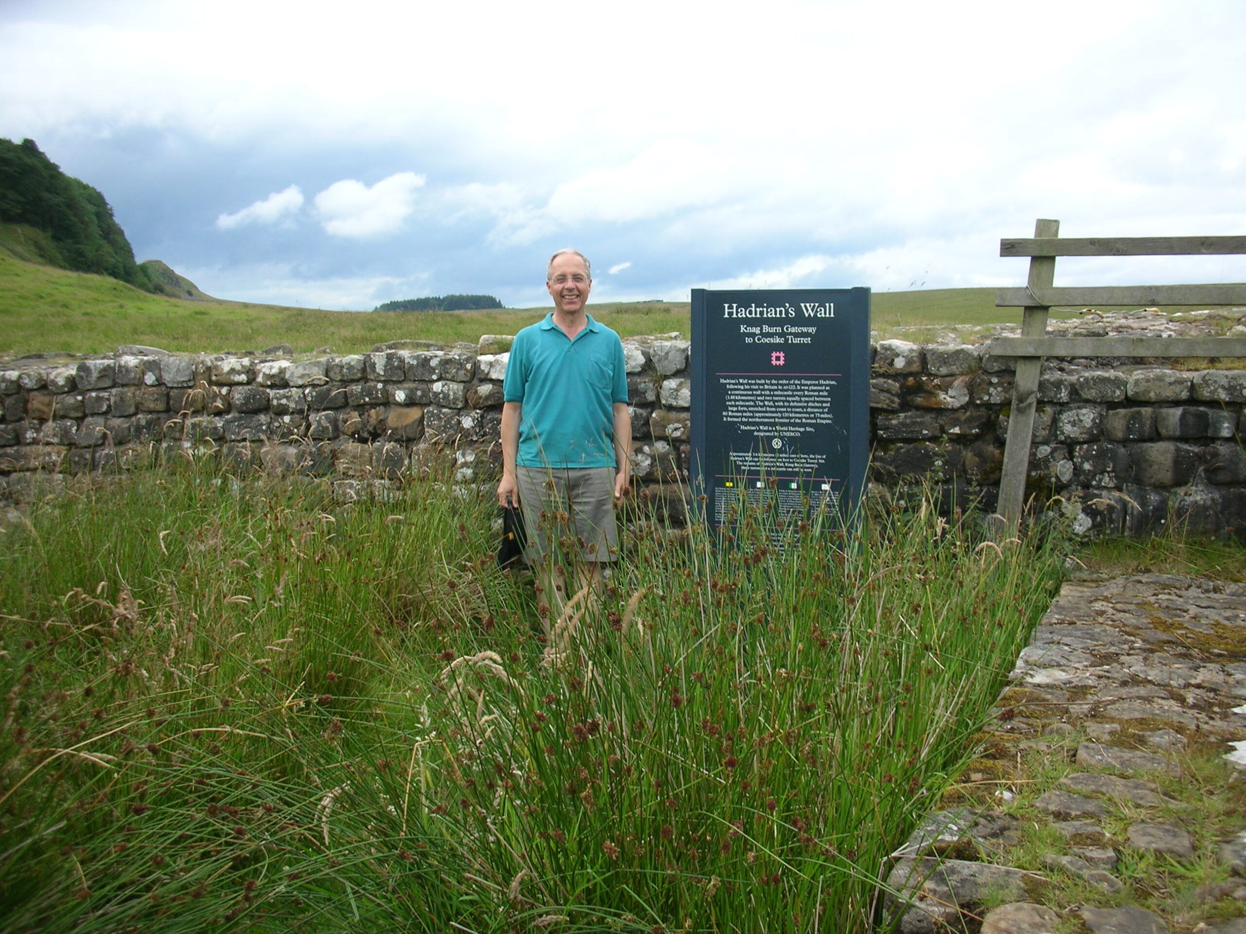 Nick smiling and standing in a green field, by Hadrian's Wall
