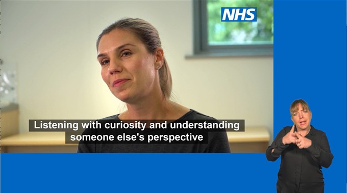 A woman is speaking in an NHS video with subtitles that read, “Listening with curiosity and understanding someone else’s perspective.” A British Sign Language interpreter is shown in bottom right corner of the screen against a blue background.