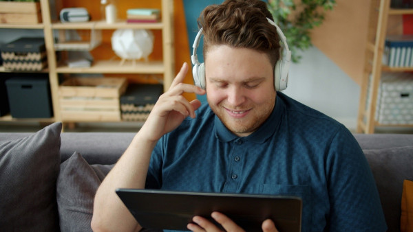 A smiling man wearing white headphones sits on a sofa, looking at a tablet. He is wearing a blue polo shirt and appears to be enjoying a video. Behind him are shelves with books, plants, and storage boxes.