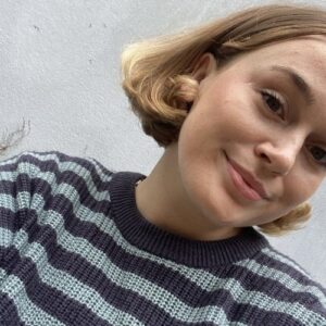 A woman with short blonde hair smiling at the camera, wearing a striped navy and light blue sweater, standing against a light grey wall.