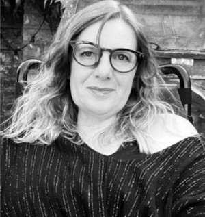 A black and white headshot of a woman with long curly hair, wearing glasses and smiling, sitting in her wheelchair, in her garden. Suzanne Bul MBE. Photo Credit - Stephane Cony