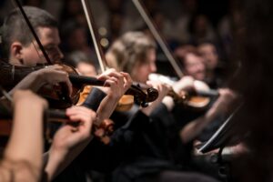 A close-up view of violinists playing in an orchestra. Several musicians are shown holding their violins and bows, with hands and instruments in focus while the background fades softly.
