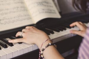 A person wearing rings and bracelets plays a piano, their hands positioned on the keys with sheet music open in front of them. The focus is on their hands and the piano keys.