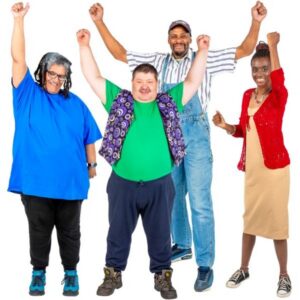 A group of four individuals standing side by side against a white background, each raising one arm in the air. They wear brightly coloured casual clothing, including a blue shirt, a patterned vest over a green top, jeans, boots, and a red cardigan over a light dress.