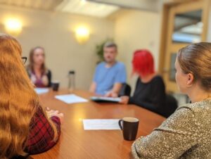 The team at A2i sit at a wooden meeting room table with papers and coffee mugs on it. The room is softly lit, and the background is slightly blurred, focusing on the attention of the discussion.