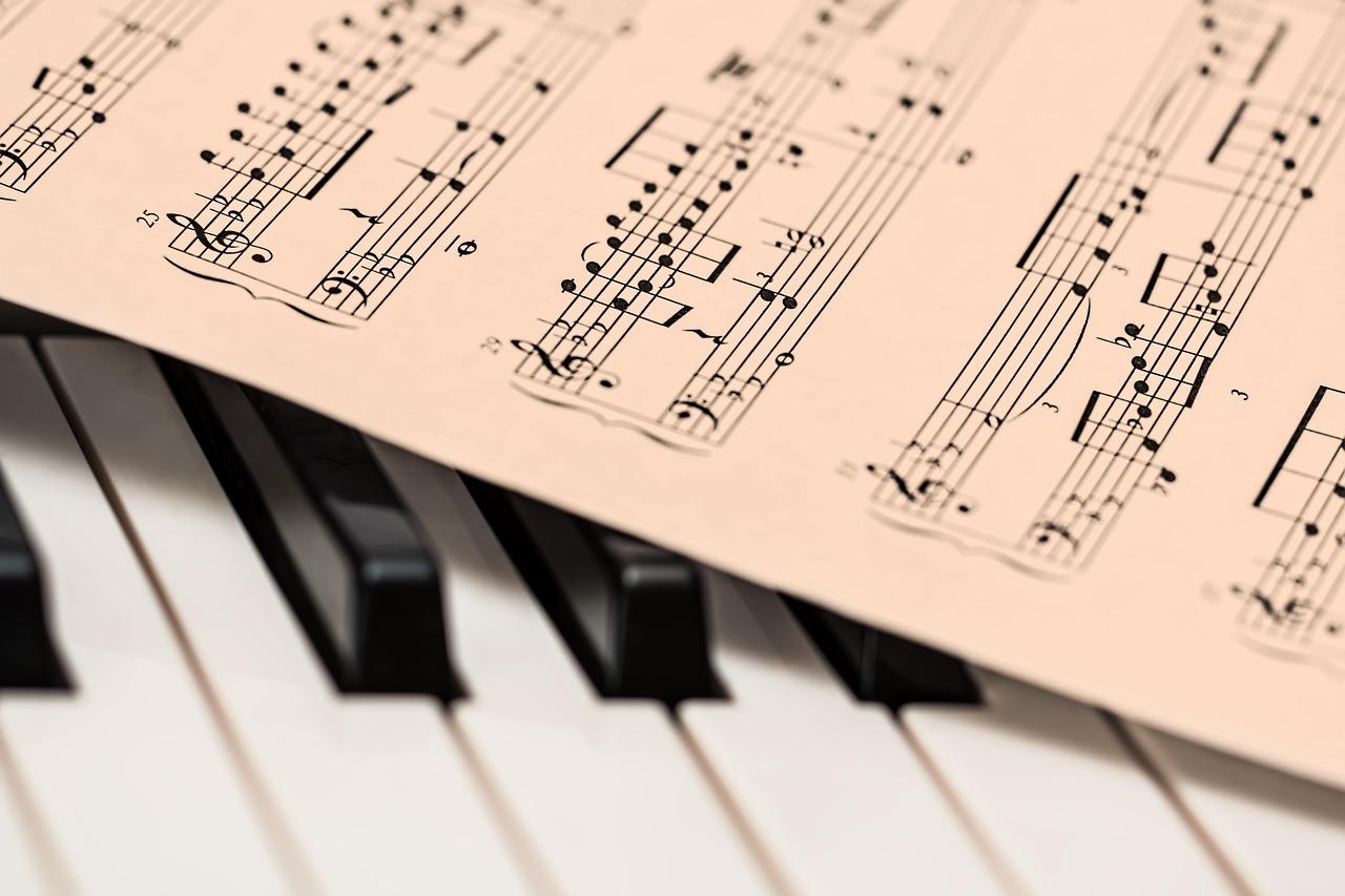 Close-up of a piano keyboard with a sheet of printed music resting across the keys. The focus is on the musical notation, with black notes and staff lines clearly visible, while the piano keys beneath are slightly out of focus.