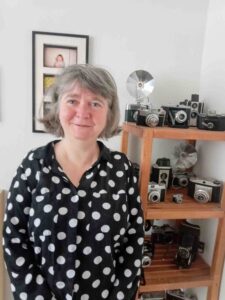 Alison stands smiling in a bright room, wearing a black blouse with large white polka dots. Behind her is a wooden shelving unit displaying a collection of vintage cameras and camera accessories, reflecting her interest in photography. A framed photograph hangs on the wall beside the shelf. The setting has a warm, personal feel and is well lit with natural light.