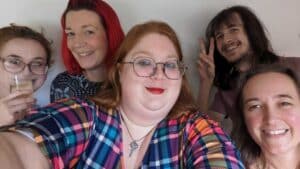 Five people smile for a group selfie. The person in the centre wears a colourful checked top, someone on the left holds a drink, and the person at the back makes a peace sign.